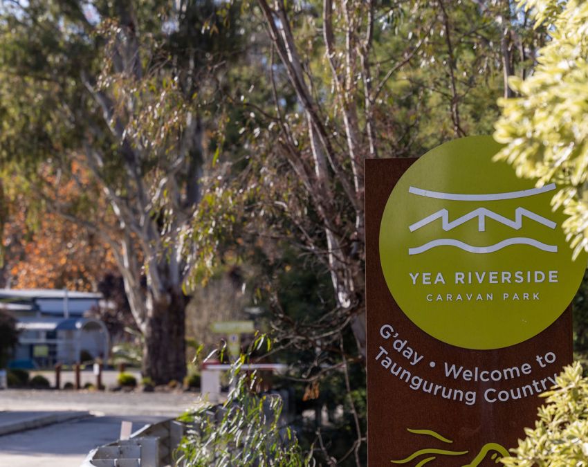 A long  view looking own a bridge with the Yea Riverside Caravan Park front entrance and Welcome Sign in the foreground