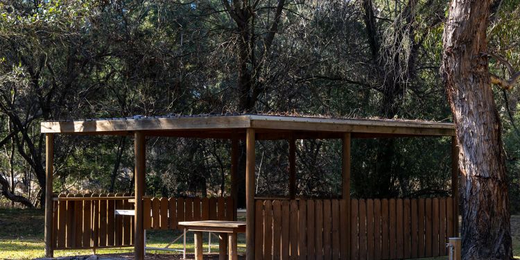 A view across a lawn of a woodens helter with wooden tables and bench seats and a low stone circular fire pit in front.
